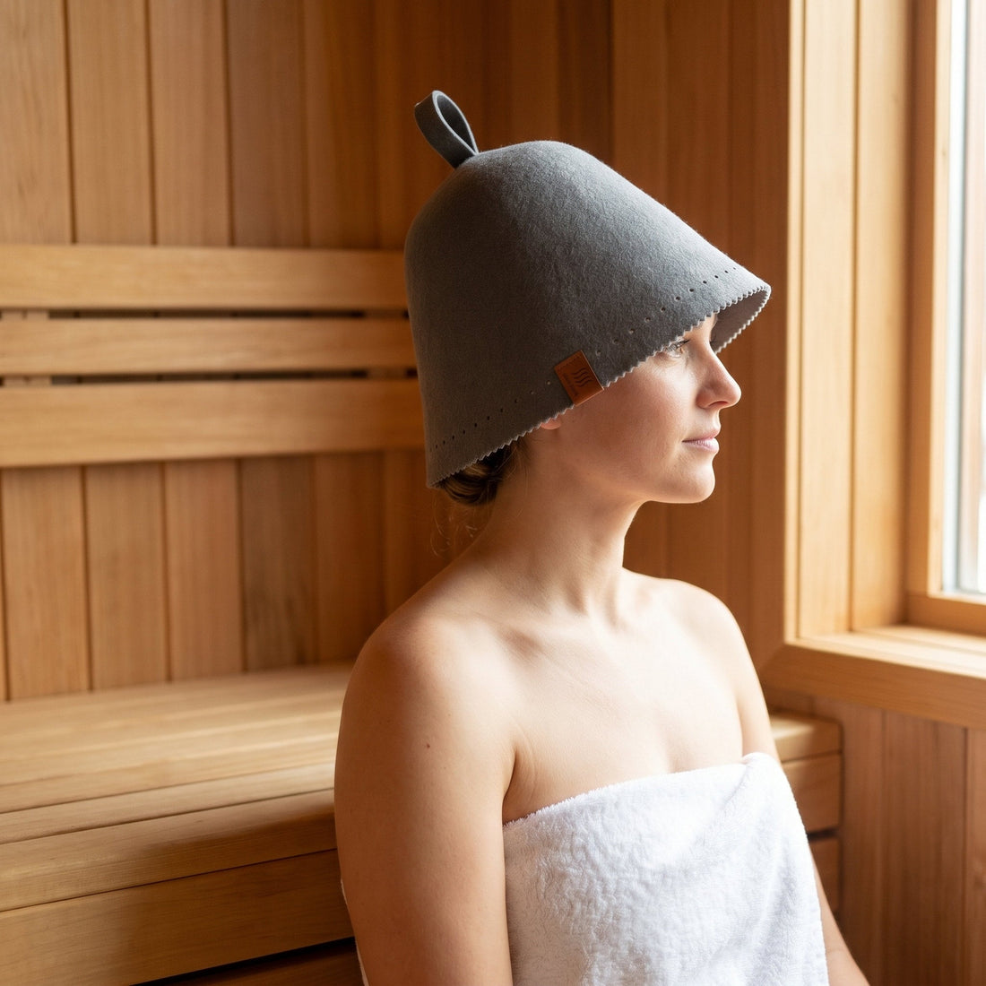 Woman in a white towel wearing a gray merino wool sauna hat by a cedar sauna window